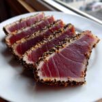 Four slices of perfectly seared ahi tuna, crusted with black and white sesame seeds, resting on a white plate.