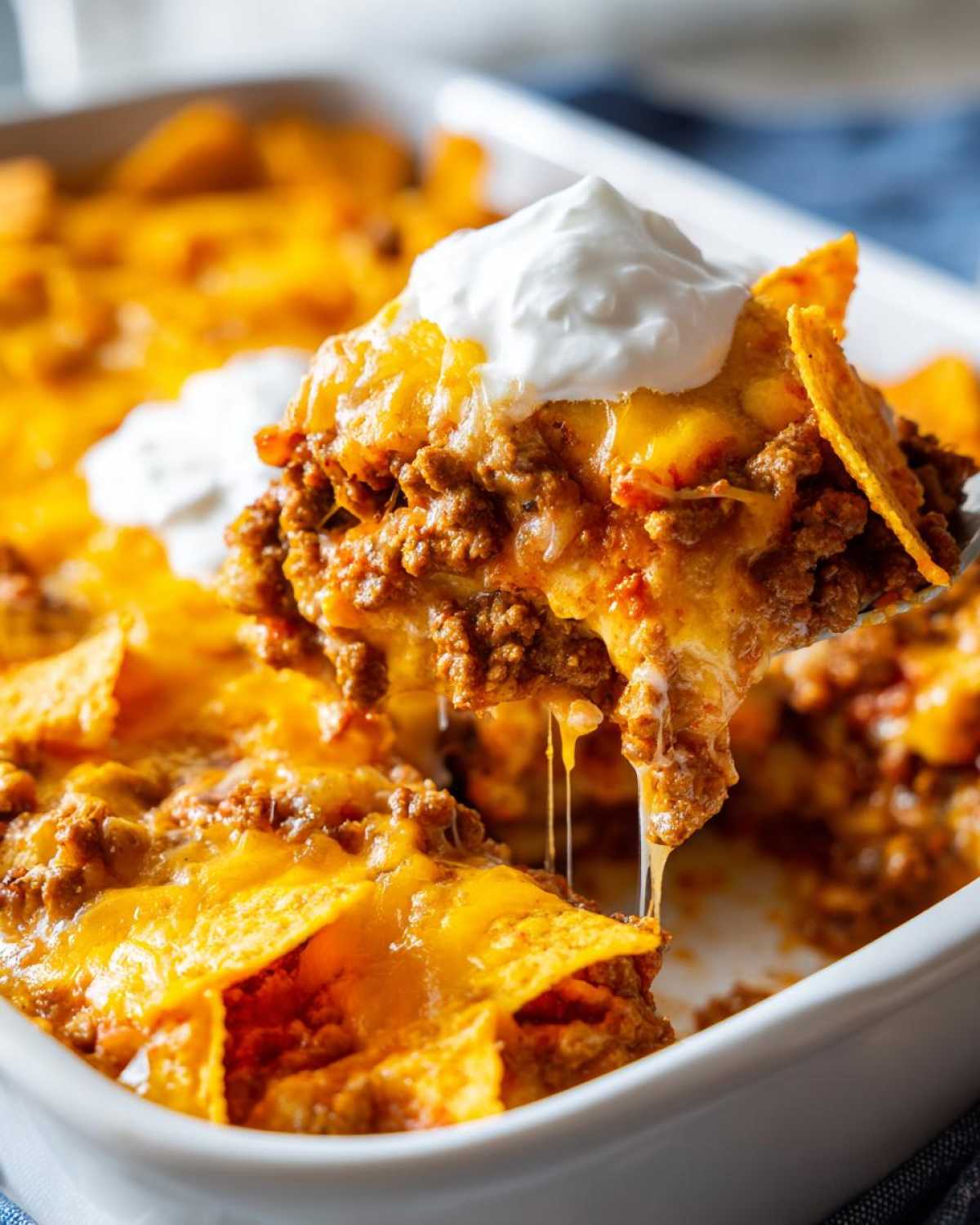 A spoonful of cheesy Dorito Casserole being lifted from a white baking dish, topped with sour cream.