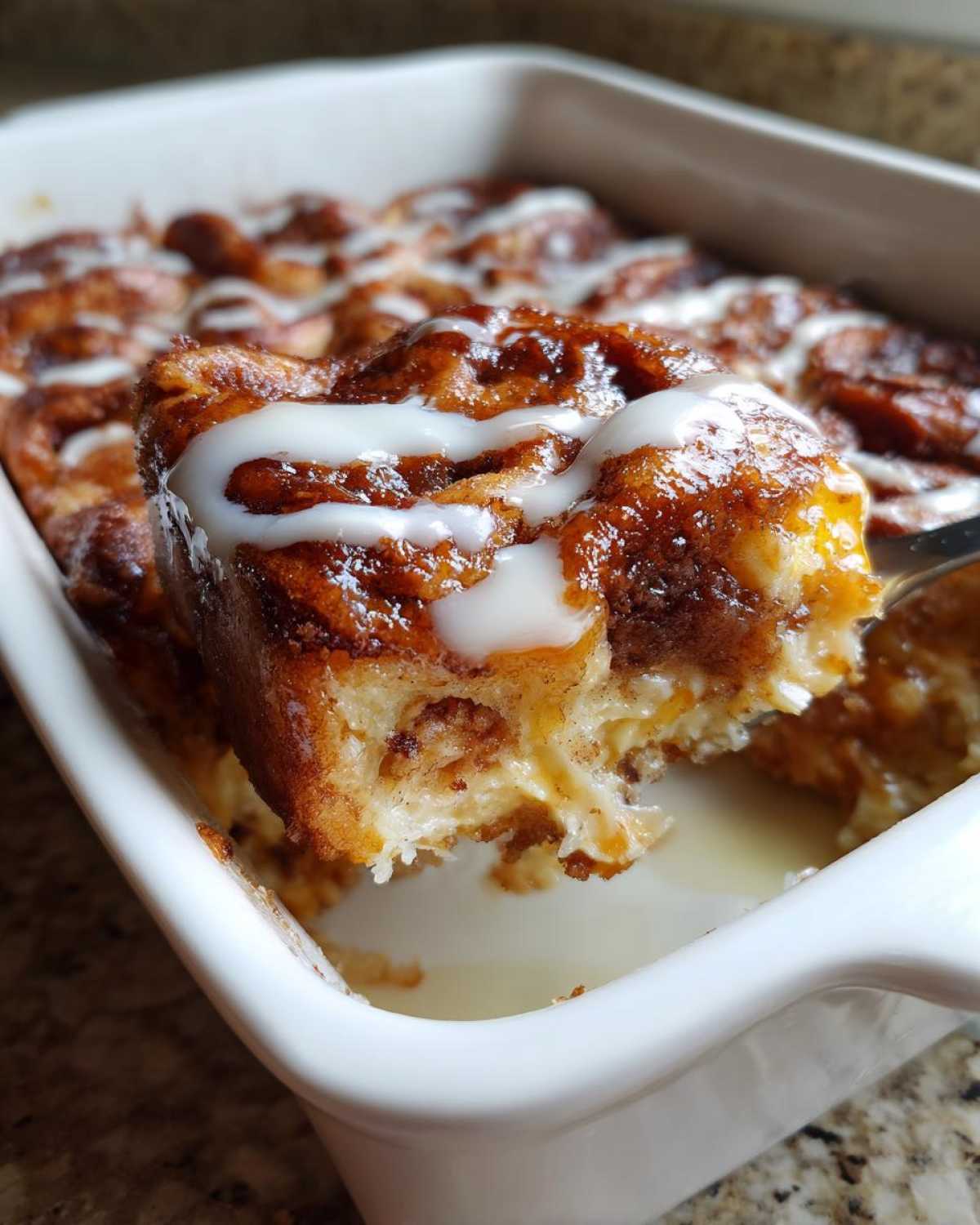 A fork lifts a gooey, iced piece of Cinnamon roll casserole from a white baking dish.