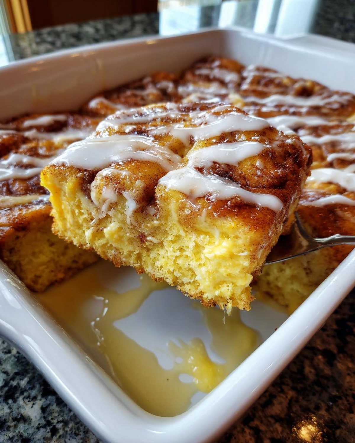 A serving spoon lifts a square piece of moist cinnamon roll casserole topped with white icing.