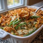 A spoonful of creamy green bean casserole topped with crispy fried onions being lifted from a white baking dish.
