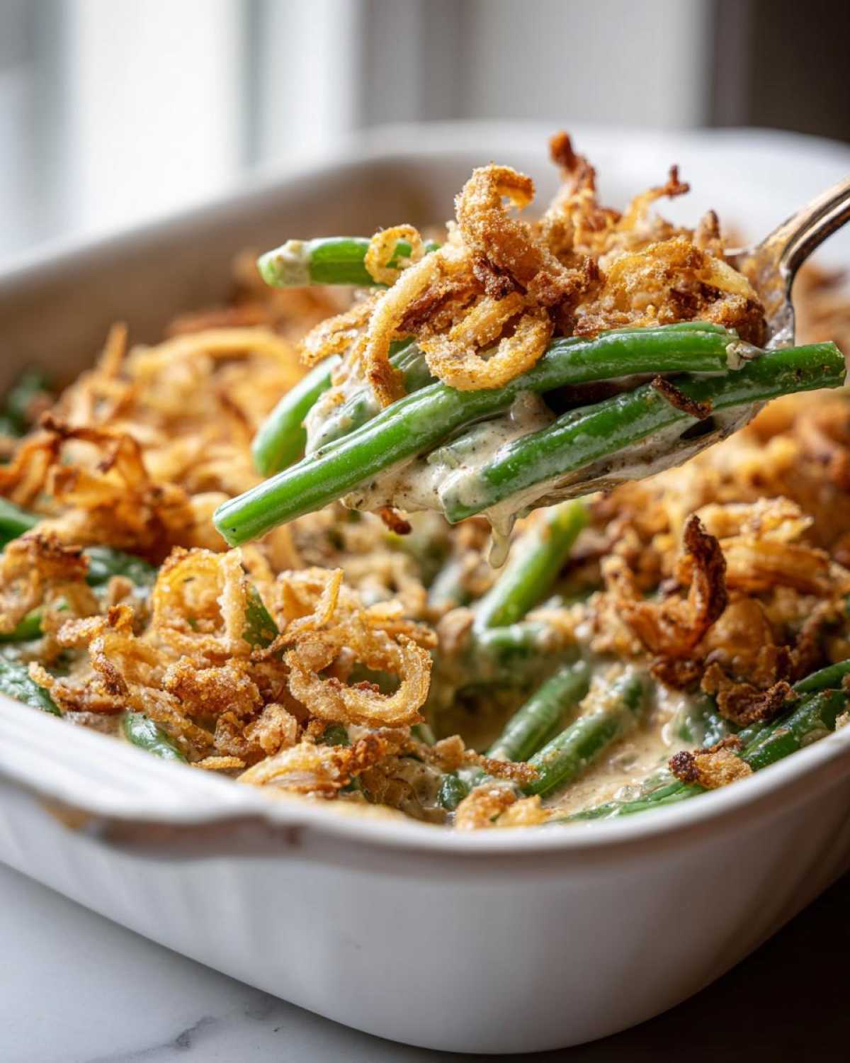 A spoonful of creamy green bean casserole topped with crispy fried onions being lifted from a white baking dish.
