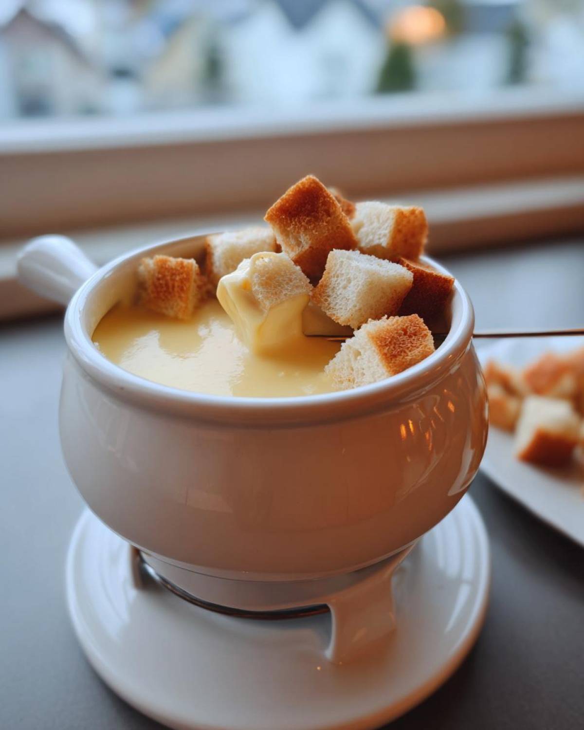 Close-up of a white pot of silky cheese fondue with toasted bread cubes being dipped.