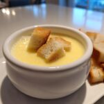 A white bowl filled with creamy, yellow cheese fondue, topped with croutons, served alongside more bread cubes.