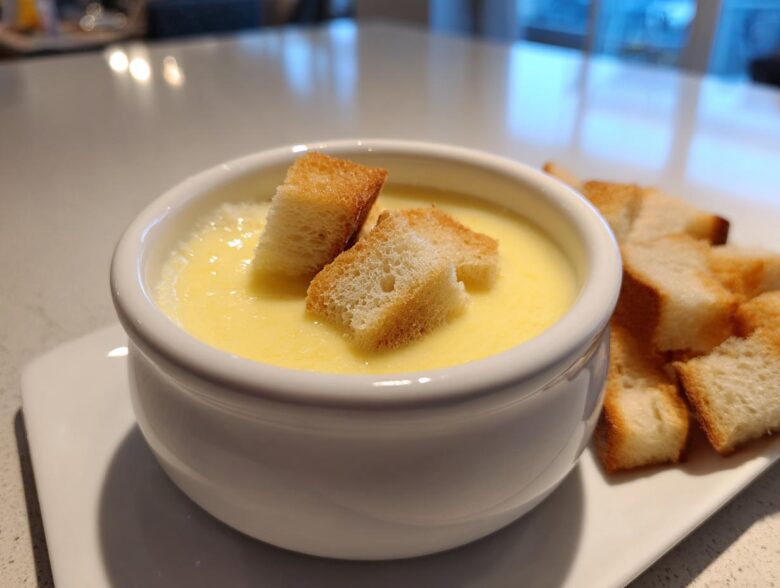 A white bowl filled with creamy, yellow cheese fondue, topped with croutons, served alongside more bread cubes.