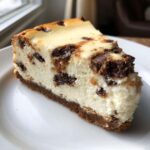 A close-up of a thick slice of creamy chocolate chip cheesecake on a white plate.