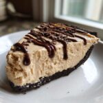 A decadent slice of no-bake peanut butter pie with a dark crust, topped with chocolate syrup and shavings.