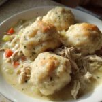 A close-up of a white bowl filled with creamy slow cooker chicken and dumplings, topped with herbs.