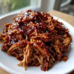 Close-up of shredded, saucy Slow Cooker Pulled Pork piled high on a white plate.