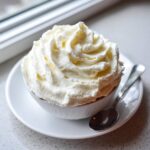 A close-up of a small white bowl topped with a generous swirl of smooth, fluffy cream cheese frosting.