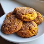 A close-up shot of several soft pumpkin snickerdoodle cookies coated in cinnamon sugar, stacked on a white plate.