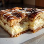 A close-up of a square slice of sopapilla cheesecake showing the thick cream cheese layer and cinnamon topping with white icing drizzle.
