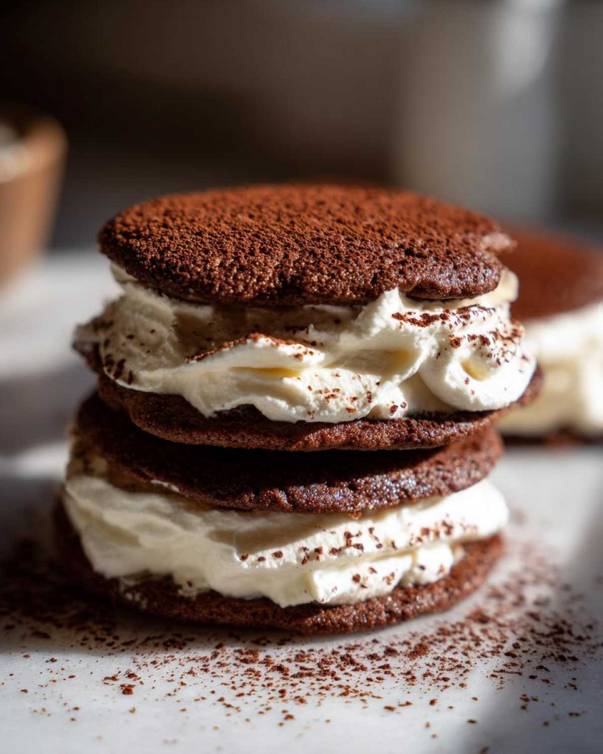 Close-up of two stacked tiramisu cookies filled with white cream and dusted heavily with cocoa powder.