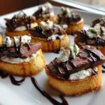Close-up of several steak crostini bites topped with cheese and balsamic glaze on a white platter.