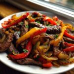 A close-up of sizzling steak fajitas featuring strips of seasoned beef, red and yellow bell peppers, and caramelized onions on a white plate.