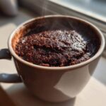 Close-up of a rich, dark chocolate mug cake steaming slightly, sitting in a light-colored mug near a window.