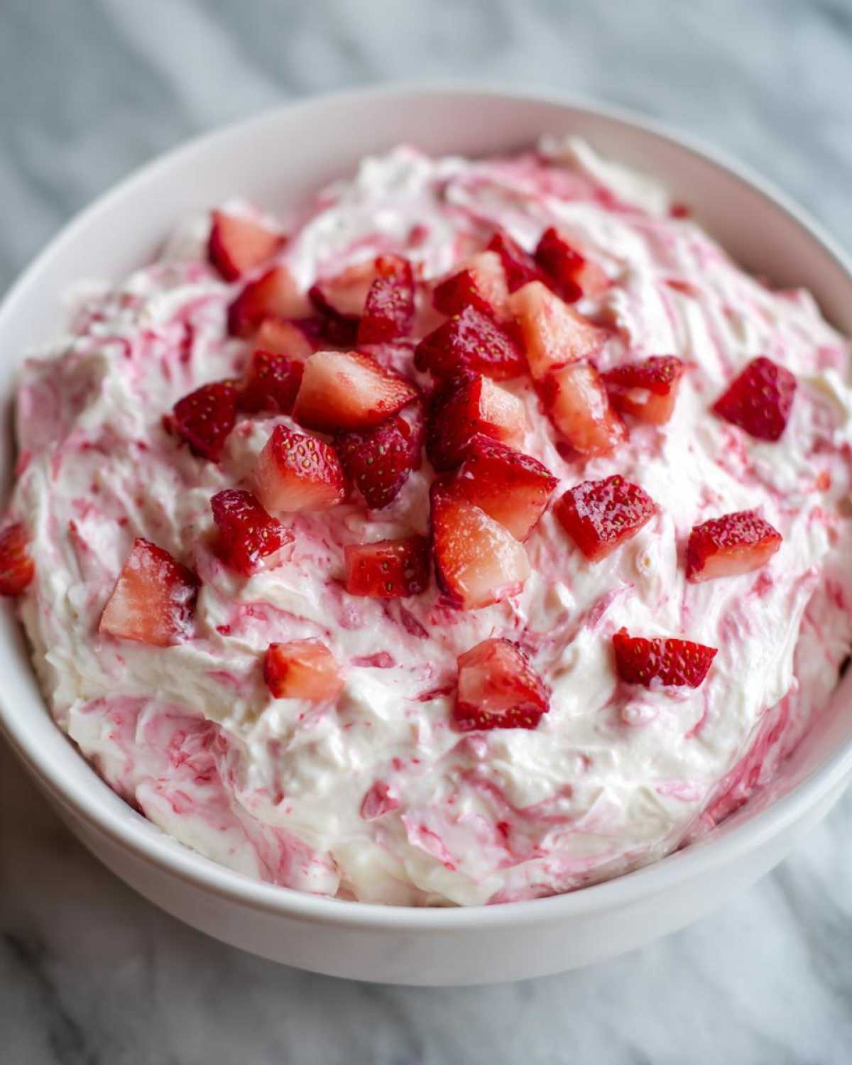 A white bowl filled with creamy, swirled strawberry cheesecake dip topped with fresh diced strawberries.