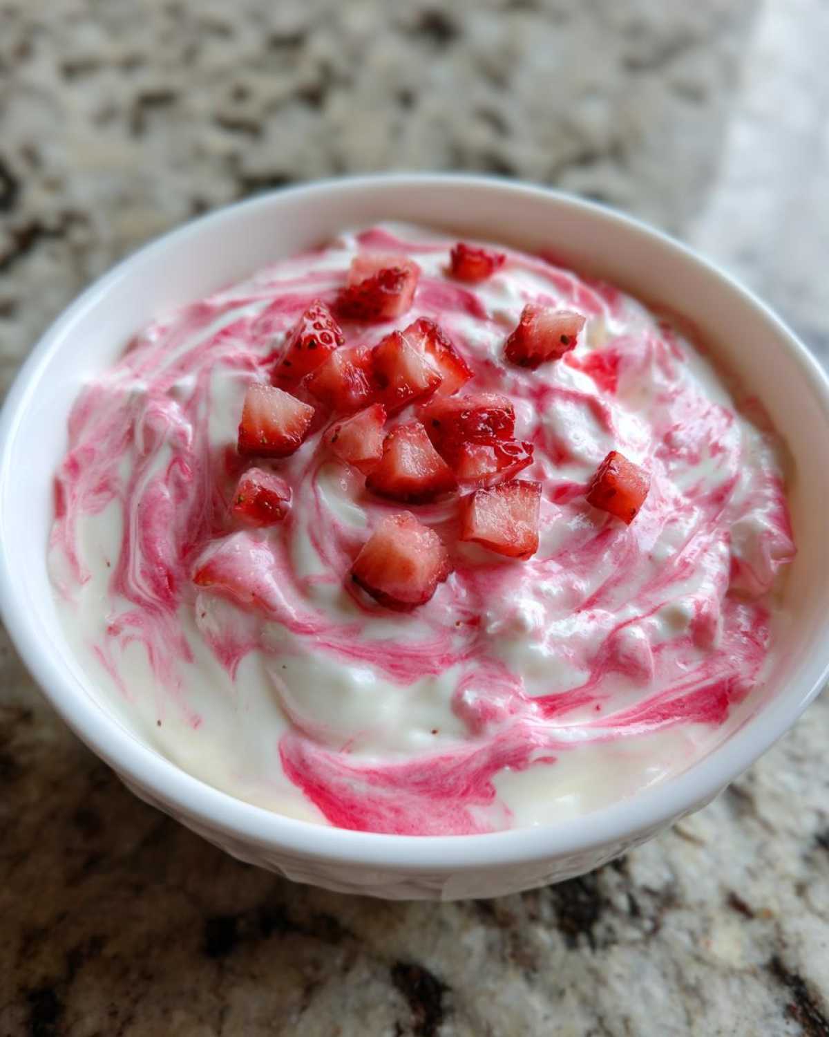 A white bowl filled with creamy strawberry cheesecake dip, swirled with pink sauce and topped with diced fresh strawberries.