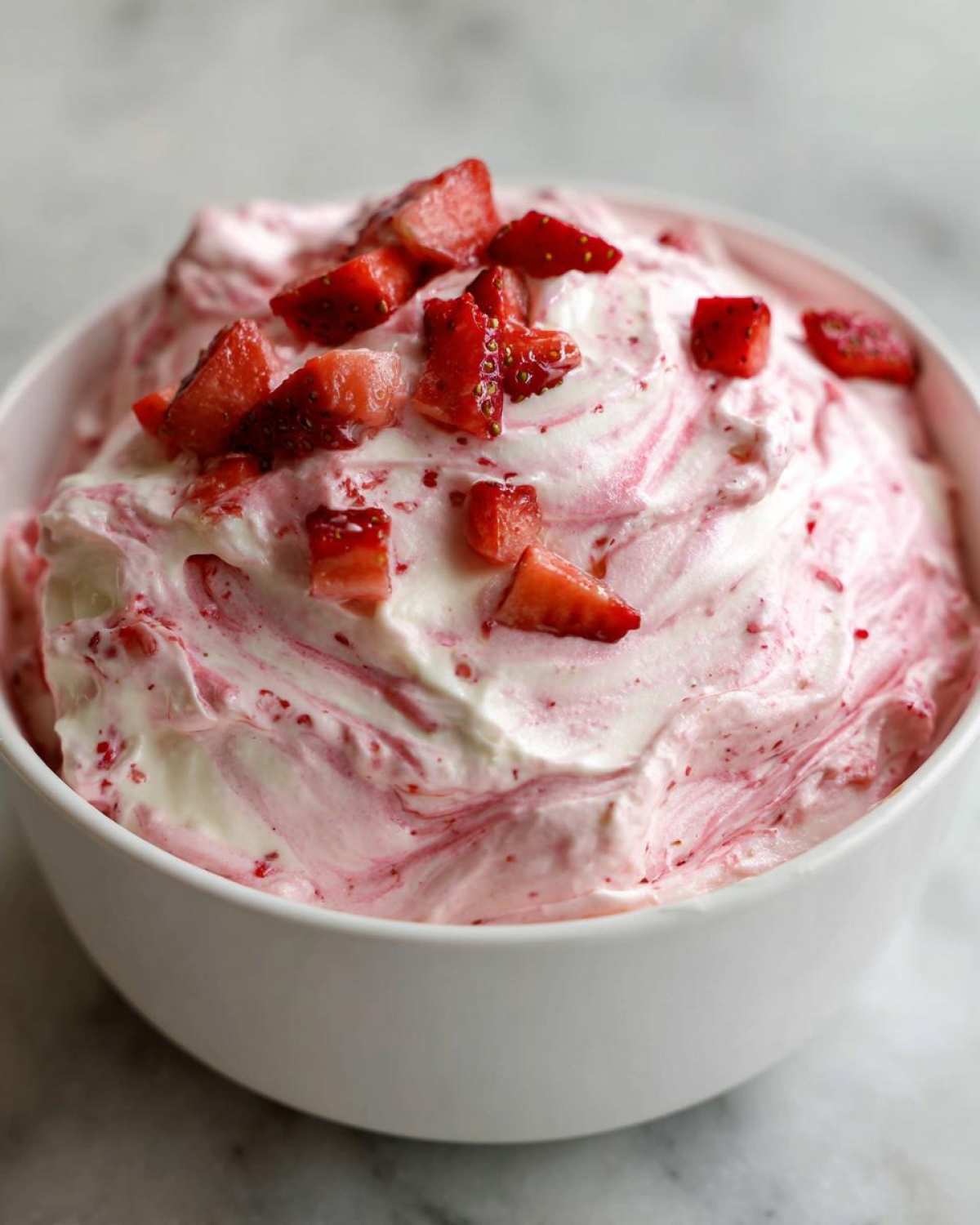 A white bowl filled with creamy, swirled strawberry cheesecake dip topped with diced fresh strawberries.