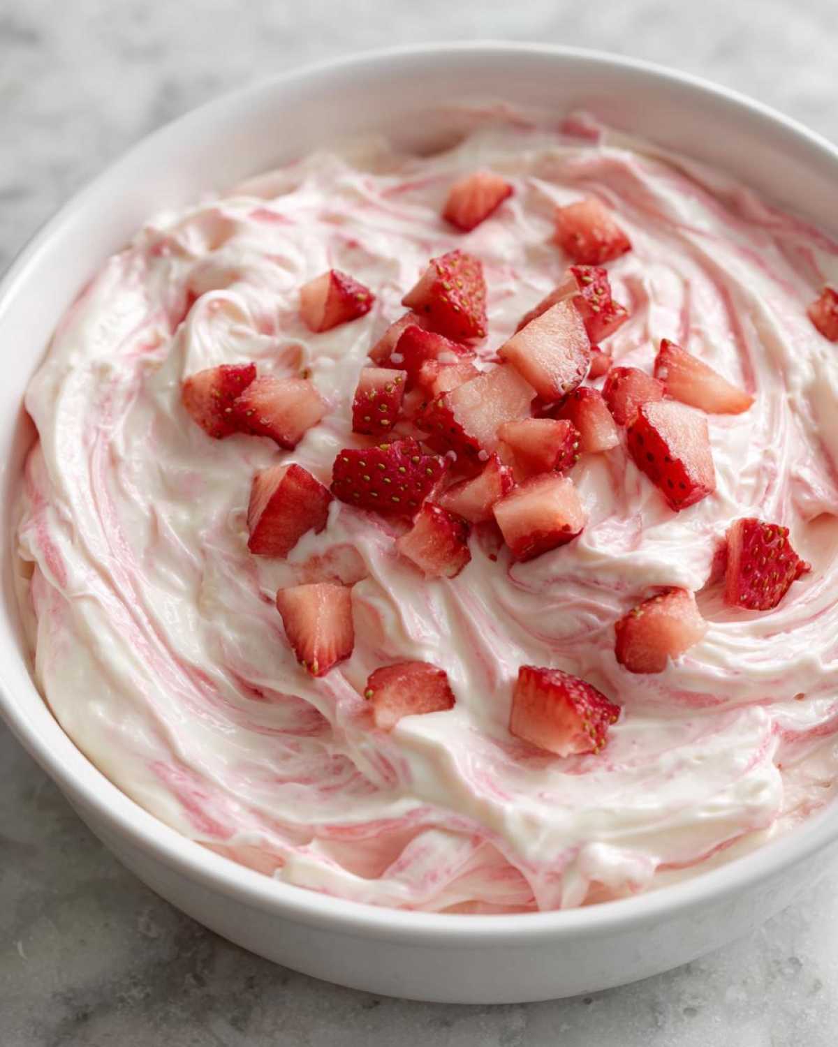 A close-up view of creamy strawberry cheesecake dip swirled with pink puree and topped with fresh diced strawberries.