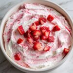 A close-up overhead view of creamy strawberry cheesecake dip swirled with pink and topped with fresh diced strawberries.