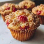 A close-up of a delicious strawberry muffin topped with brown sugar crumble, with more strawberry muffins blurred in the background.
