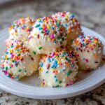 A stack of white chocolate-coated sugar cookie truffles generously covered in colorful rainbow sprinkles on a white plate.