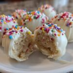 A close-up view of several white chocolate coated sugar cookie truffles, one of which is broken open to show the cookie dough center.