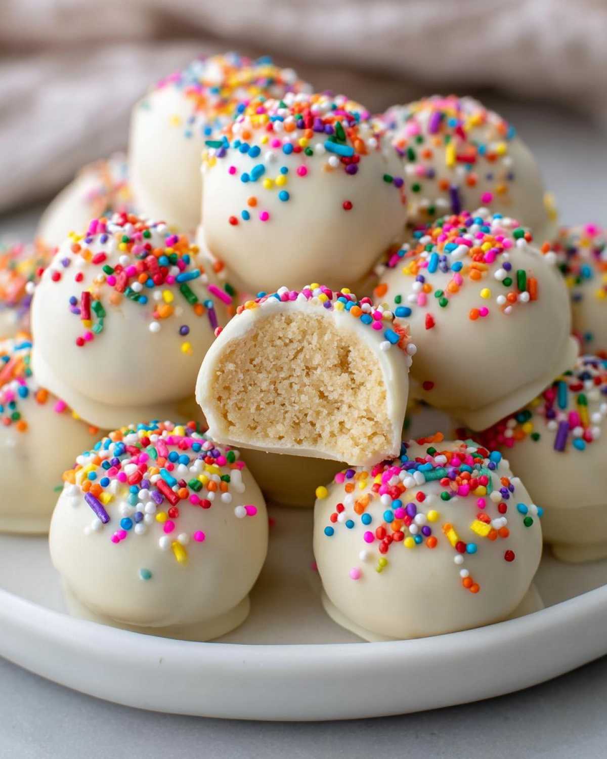 A pile of white chocolate-covered sugar cookie truffles topped with colorful sprinkles, one cut in half showing the soft interior.