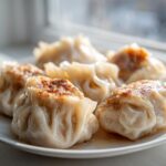 A close-up of several pan-fried Traditional Chinese Dumplings with golden-brown bottoms resting on a white plate.