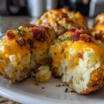 A close-up view of a twice baked potato split open, showing fluffy interior topped with melted cheddar cheese, bacon bits, and chives.