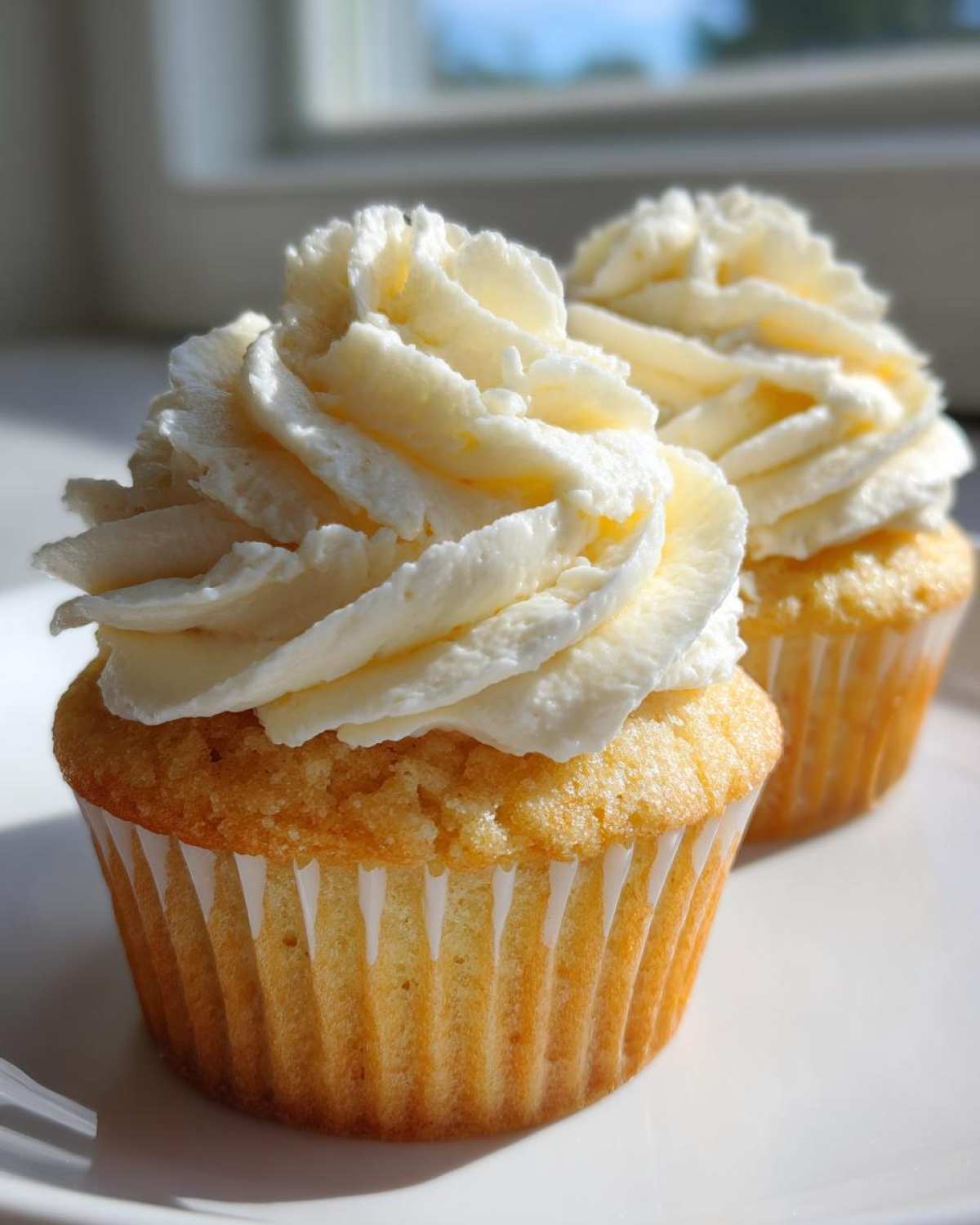 Two perfectly baked vanilla cupcakes topped with high swirls of white frosting, sitting on a white plate.