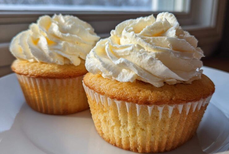 Two perfectly baked vanilla cupcakes topped with tall swirls of white cream frosting, sitting on a white plate.