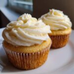 Close-up of two moist vanilla cupcakes topped with swirls of white vanilla frosting on a white plate.