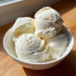 Four scoops of creamy vanilla protein ice cream served in a white bowl, highlighted by bright sunlight.
