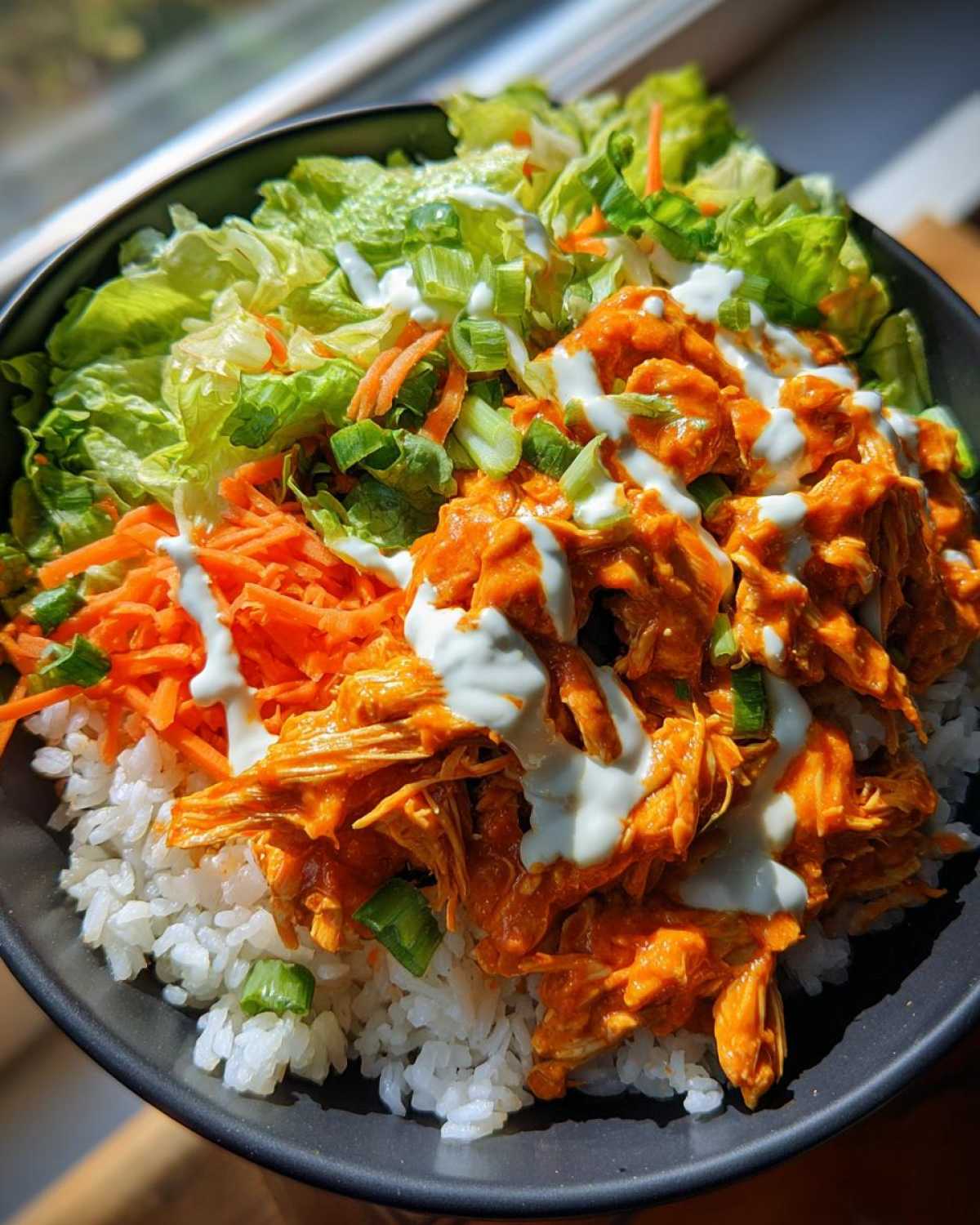 Close-up of a delicious buffalo chicken bowl featuring shredded orange buffalo chicken over white rice, topped with lettuce, carrots, and ranch dressing.