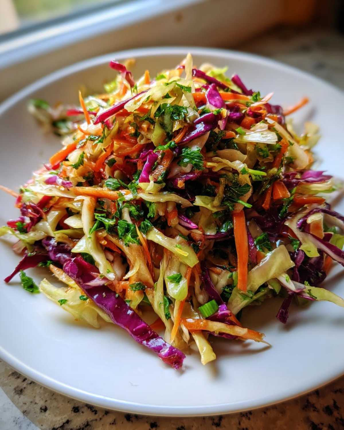 A close-up of a colorful, fresh cabbage salad featuring shredded green and red cabbage, carrots, and green herbs on a white plate.