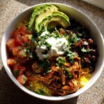A vibrant, overhead view of a delicious chicken burrito bowl featuring shredded chicken, yellow rice, black beans, salsa, avocado, and sour cream.