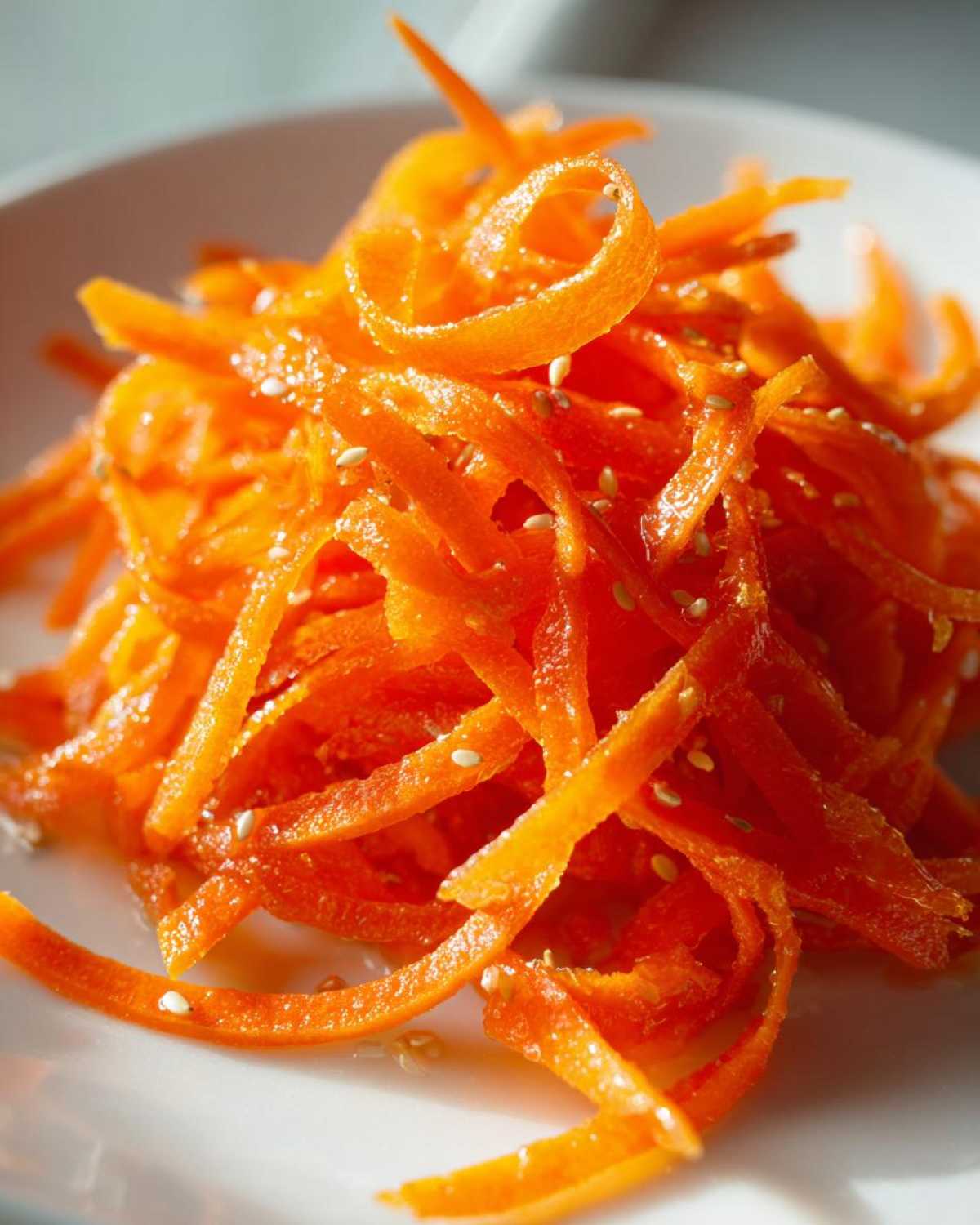 Close-up of a bright orange mound of raw carrot salad, thinly shaved and glistening, topped with white sesame seeds.
