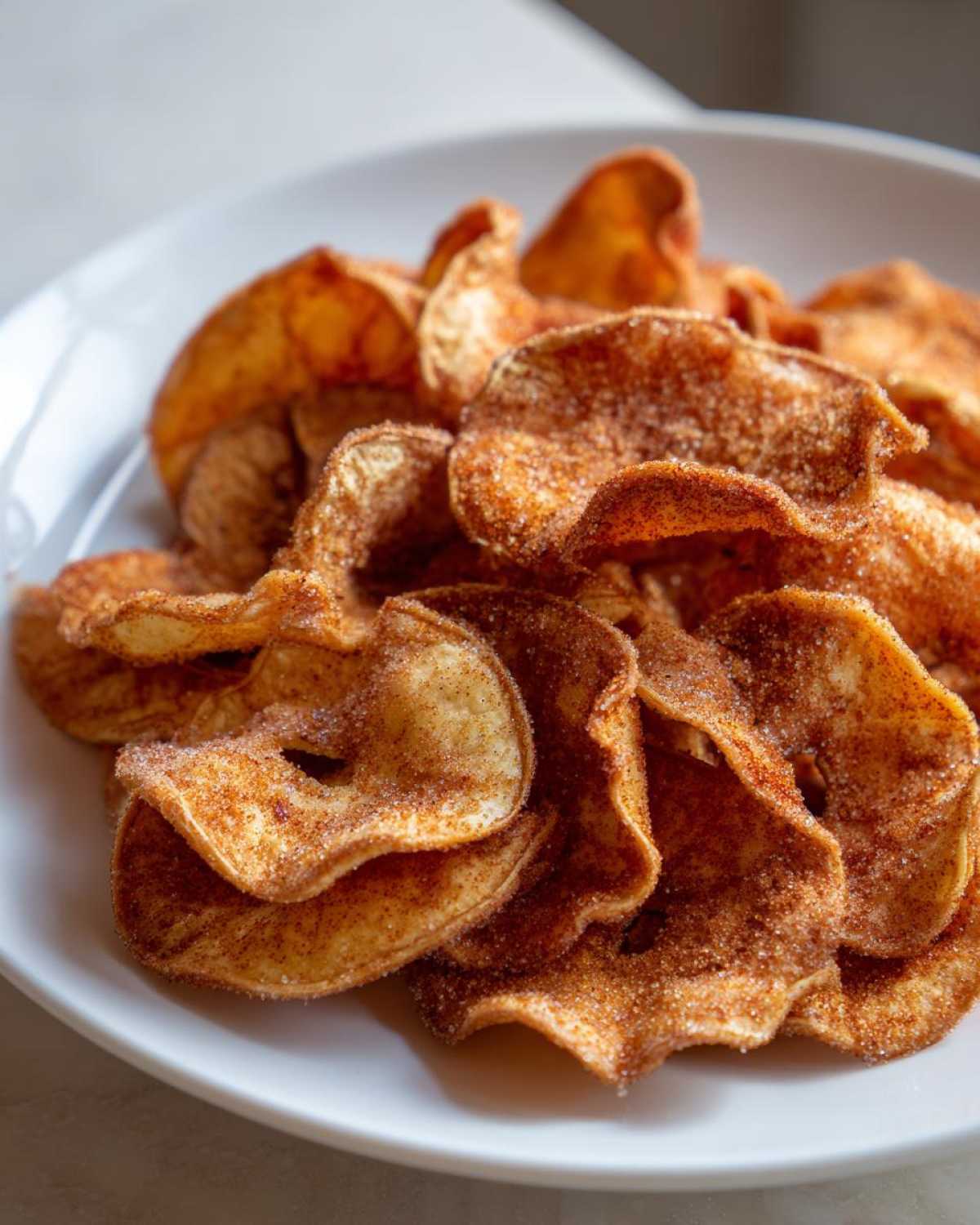 Close-up of crispy Air Fryer Cinnamon Sugar Apple Chips heavily coated in cinnamon sugar, piled on a white plate.