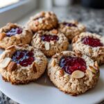 Close-up of several Gluten-Free Raspberry Almond Thumbprint Cookies topped with bright red jam and sliced almonds.