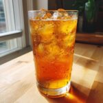 A tall glass filled with condensation and ice, holding refreshing Iced Peach Green Tea, sitting on a wooden table.