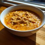 A close-up of a white bowl filled with rich, orange Instant Pot Creamy Chicken Taco Soup topped with shredded chicken and corn.