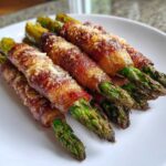 Close-up of crispy Keto Bacon Wrapped Asparagus Bundles topped with melted Parmesan cheese on a white plate.