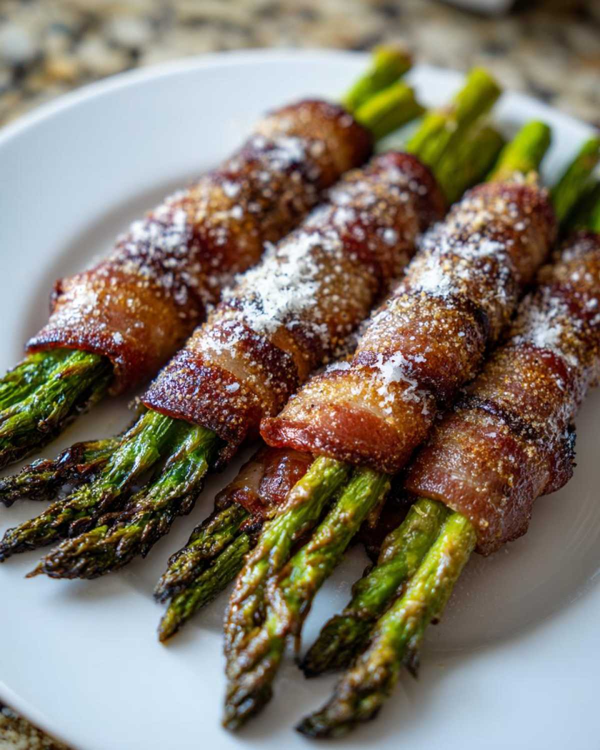 Close-up of four Keto Bacon Wrapped Asparagus Bundles, crispy bacon topped with grated Parmesan cheese, served on a white plate.