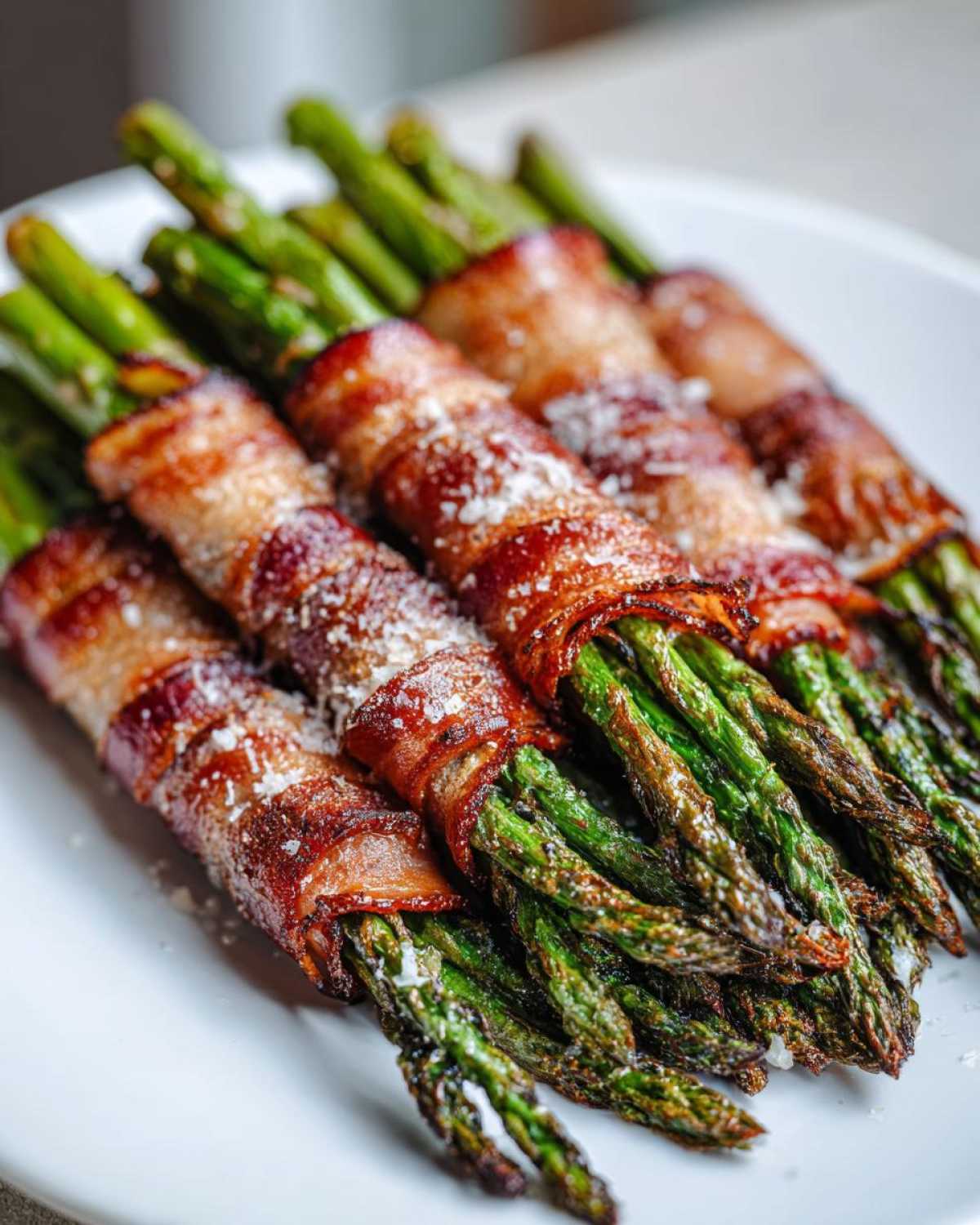 Close-up of perfectly cooked Keto Bacon Wrapped Asparagus Bundles sprinkled with Parmesan cheese on a white plate.
