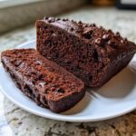 A loaf and a slice of moist Gluten-Free Chocolate Zucchini Bread topped with chocolate chips on a white plate.