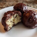 Three No-Bake Chocolate Chip Cookie Dough Truffles on a white plate, one bitten to show the doughy interior.