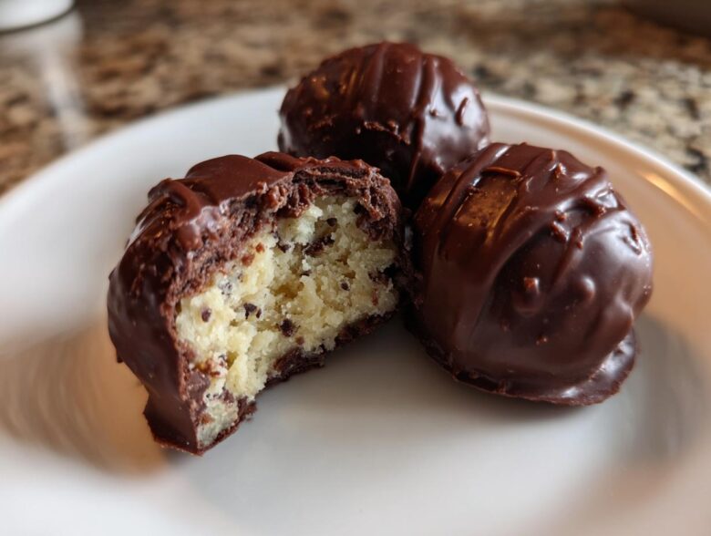 Three No-Bake Chocolate Chip Cookie Dough Truffles on a white plate, one bitten to show the doughy interior.