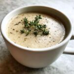 Close-up of a white mug filled with Vegan Creamy Mushroom Soup with Thyme, garnished with fresh thyme sprigs.